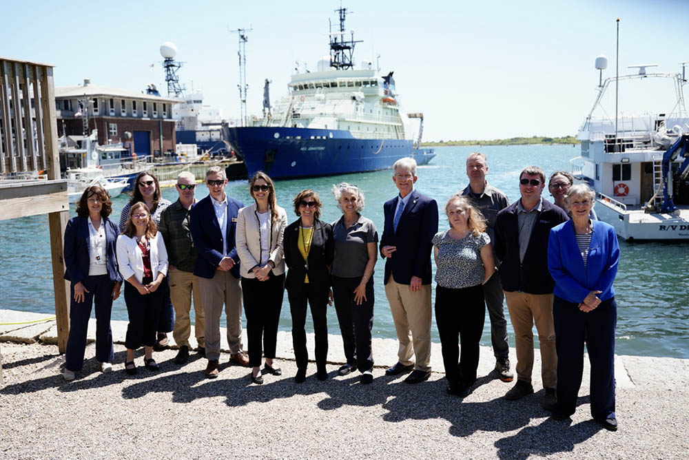 A group of policymakers and scientists stands by the waterfront with boats in the background.