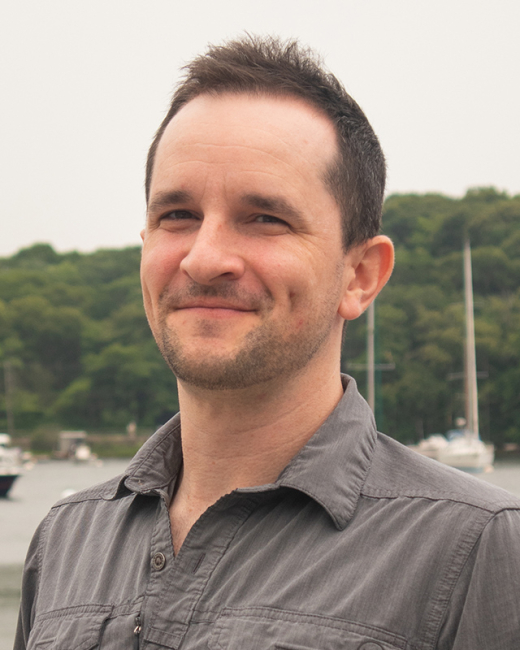 Male presenting person with short brown hair standing in front of the water.
