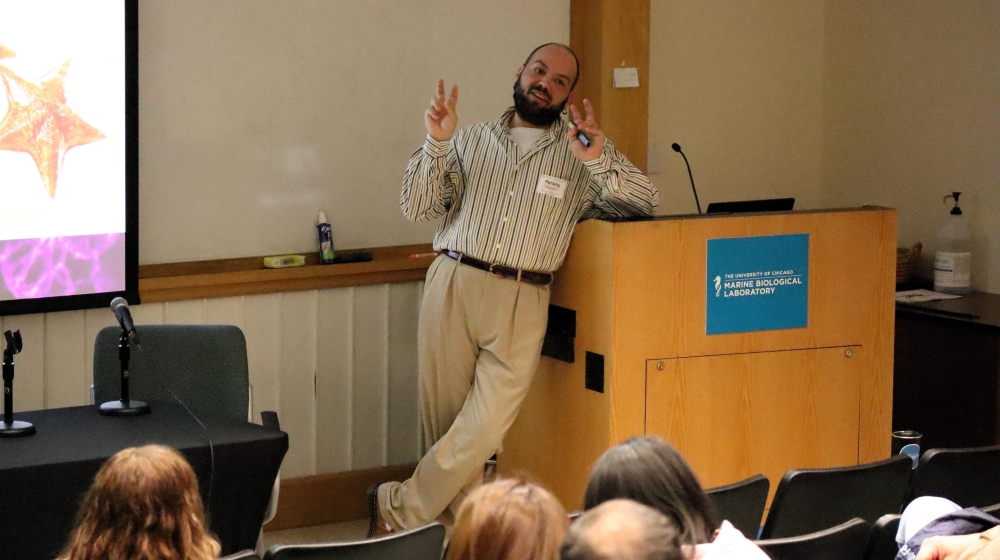 A man leads against a lectern and addresses viewers in an auditorium. He is making the "air quotes" sign.