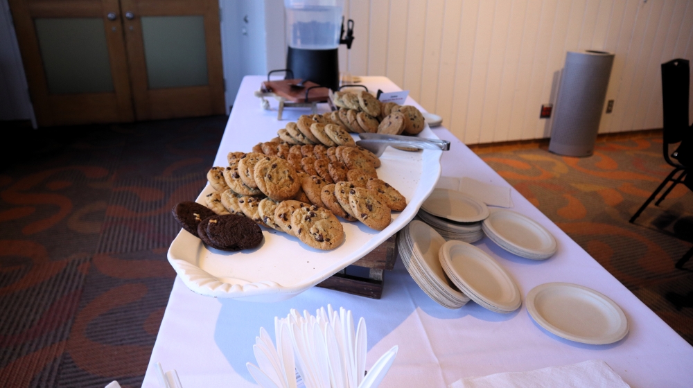 Various types of cookies sit on a white tray. Also on the table are disposable plates and cutlery and a drink container.