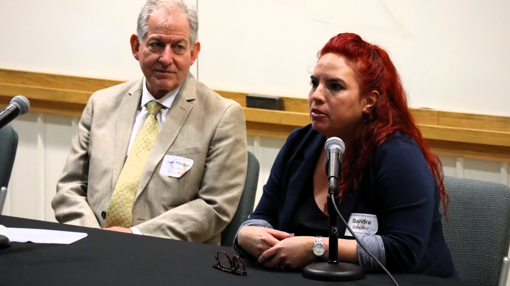 A man in a beige suit and a woman wearing navy blue sit at a table with microphones in front of them.