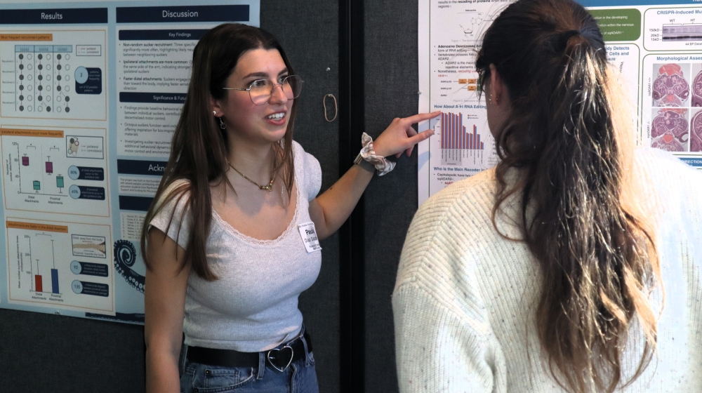 Two women stand in front of a poster on a bulletin board. The one on the left is pointing to the poster, while the one on the right faces the poster.