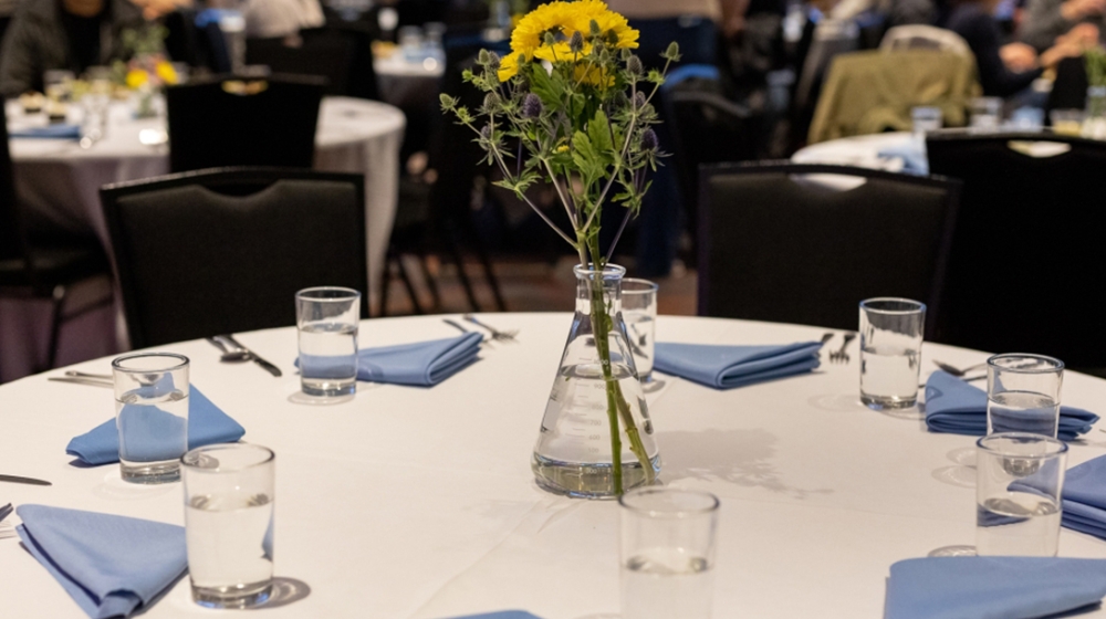 Round table with place settings and flowers, with people seated at more tables in the background.