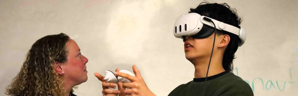 A woman and a young man stand in front of a whiteboard. The man is wearing a VR headset and holding two VR controllers in his hands.