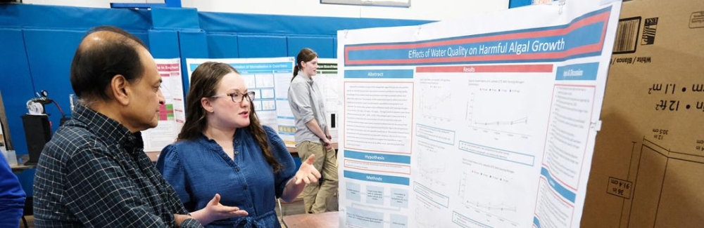 A young woman and a man stand in front of a poster set on a table.