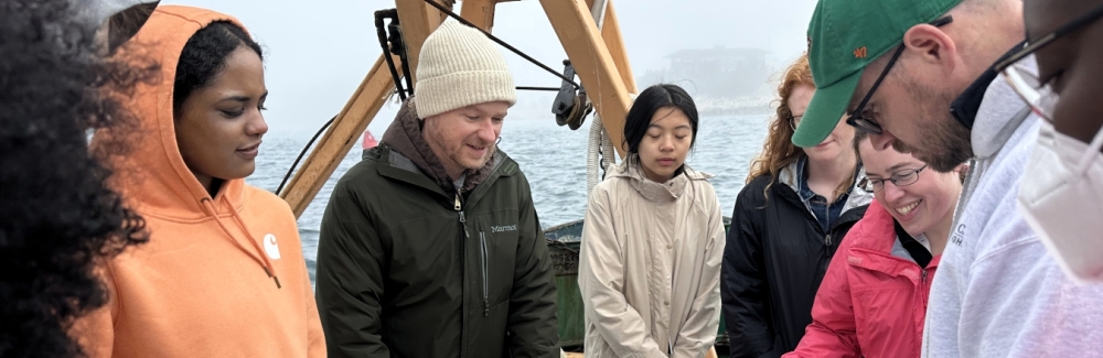 People gather in a circle around empty totes on a boat and look at a map.
