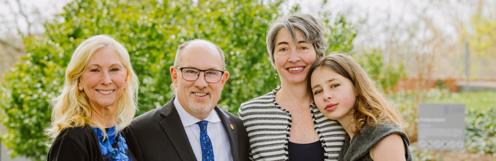 Three women and a man stand in front of some shrubbery.