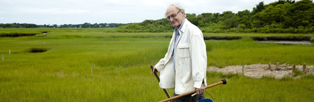 A man in a white lab coat and boots carries a bucket and other field equipment through a marsh.
