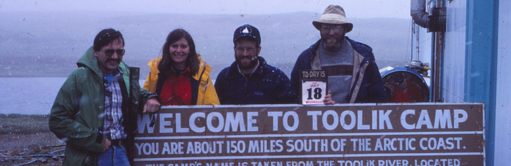 Four people pose behind/in front of a sign that says "Welcome to Toolik Camp". A wide expanse of land and water is visible behind them, and a building is next to them.