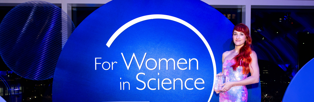 Kaitlyn Webster wearing a gown, holding an award, standing to the right of a "For Women in Science" sign. 