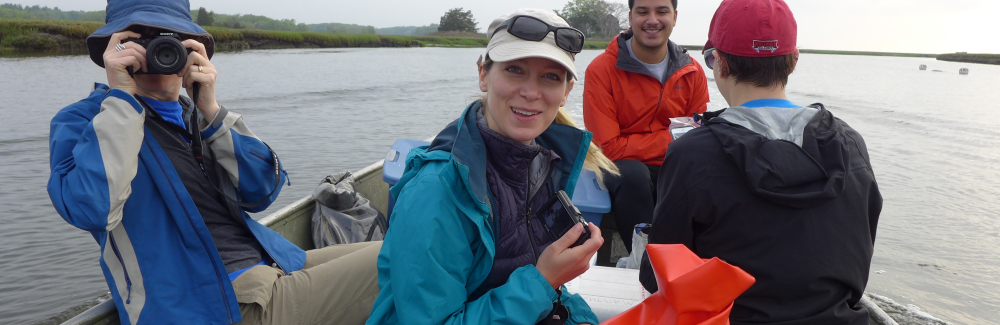 four people on a small row boat on the water.  one on left has a camera to face. one in back has a red jacket. Person in the forground has a bag and the one to the far right is facing backwards. 