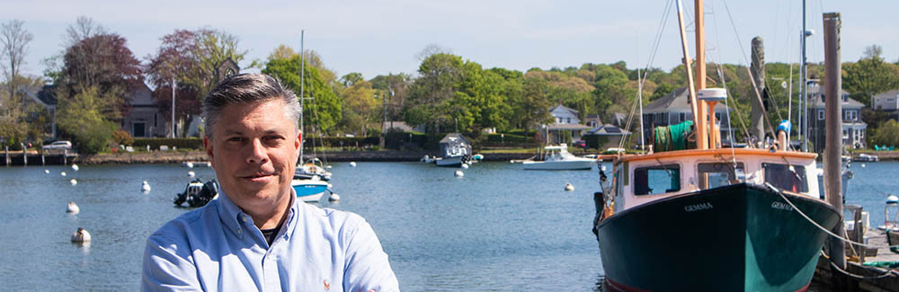 Man in a blue shirt standing and smiling on a dock with a research vessel behind him.