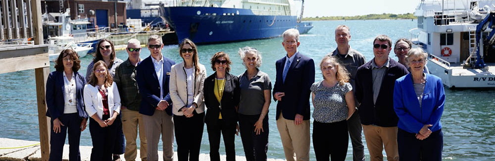 A group of policymakers and scientists stands by the waterfront with boats in the background.