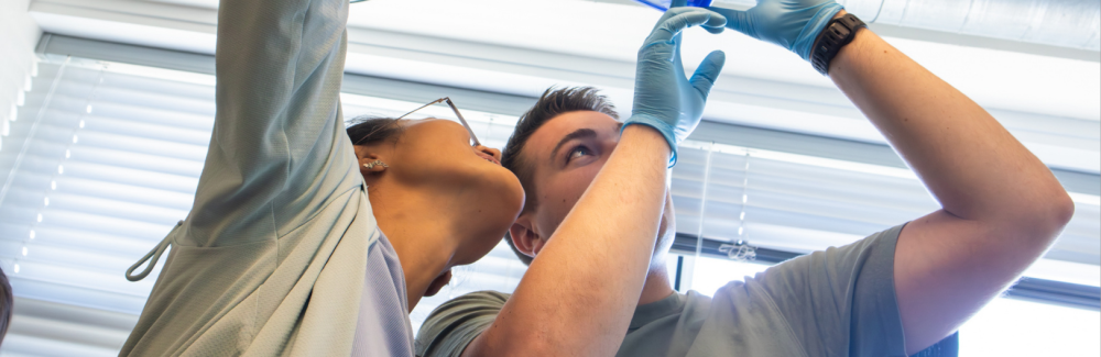 This photo shows two students holding up two containers filled with blue colored gels and looking up at them.