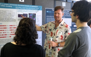 A man stands in front of a poster on a bulletin board and points at it, facing away from the board. Another man and a woman stand in front of him, facing the poster.