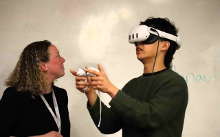 A woman and a young man stand in front of a whiteboard. The man is wearing a VR headset and holding two VR controllers in his hands.