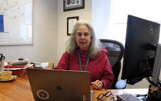A woman sits at a desk and looks at an open laptop.