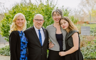 Three women and a man stand in front of some shrubbery.