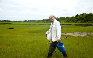A man in a white lab coat and boots carries a bucket and other field equipment through a marsh.