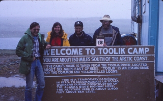 Four people pose behind/in front of a sign that says "Welcome to Toolik Camp". A wide expanse of land and water is visible behind them, and a building is next to them.