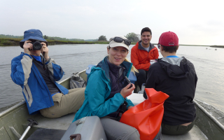 four people on a small row boat on the water.  one on left has a camera to face. one in back has a red jacket. Person in the forground has a bag and the one to the far right is facing backwards. 