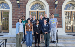 A group of researchers gathers on the steps of a brick building for a cohort photo.