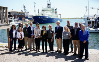 A group of policymakers and scientists stands by the waterfront with boats in the background.