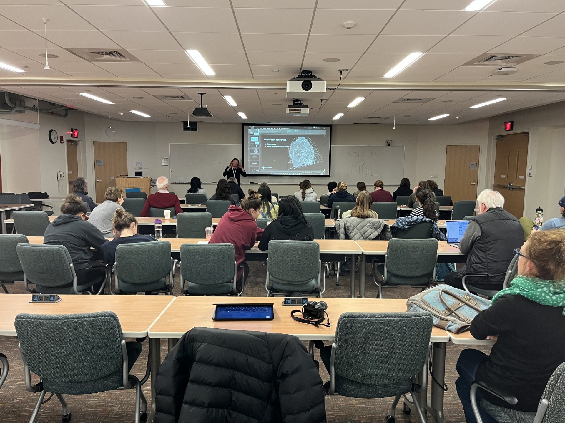 A woman stands in front of a whiteboard and projector screen at the front of a room. Rows of tables and chairs fill the rest of the shot, with people scattered throughout.