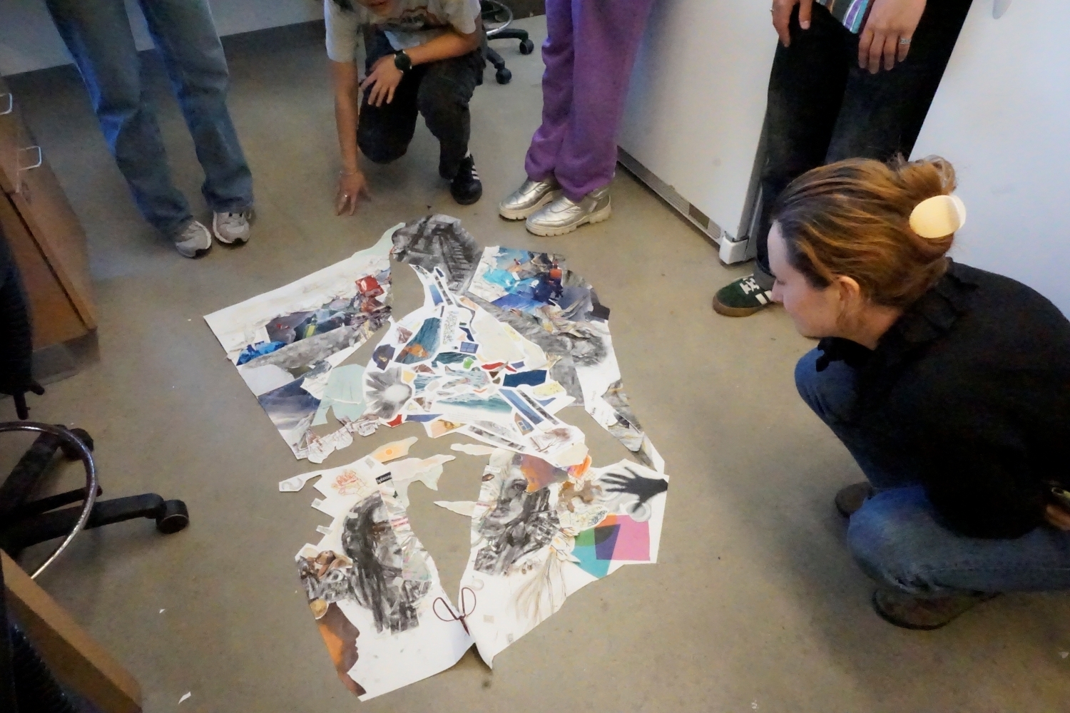 A woman crouches next to a piece of collage art laying on the floor. Other peoples' feet are visible around the artwork.