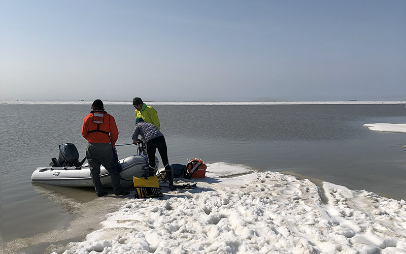 Inflatable boat up on a snowy shore with three people loading gear.