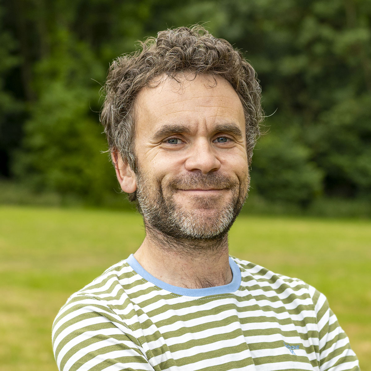 Smiling man with short curly brown hair wearing a green striped shirt standing on a lawn with trees in the background.