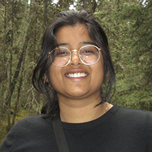 Female presenting person wearing glasses, standing in front of trees.