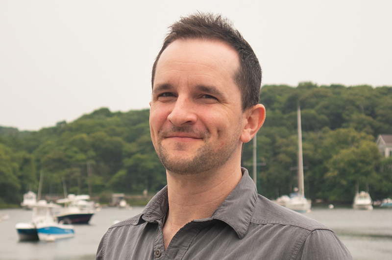 Male presenting person with short brown hair standing in front of the water, with boats in the background.