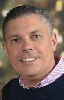 Smiling man with short dark hair wearing a pink shirt and dark sweater.