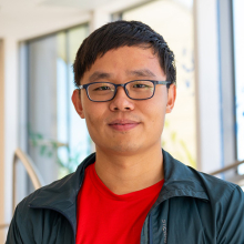 Man with dark short hair wearing glasses and a red shirt and dark jacket.