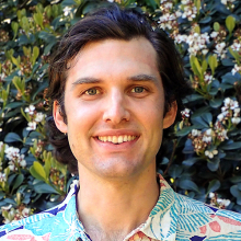 Male-presenting person with dark hair standing in front of a flowering bush.