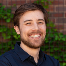 Male-presenting person with dark hair and short beard standing in front of an ivy covered brick wall.