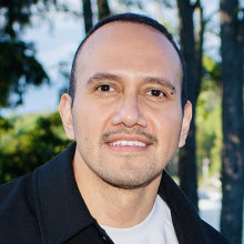 Male presenting person with short dark hair standing in front of trees.