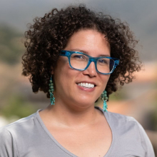 Female presenting person with curly brown hair and blue glasses.