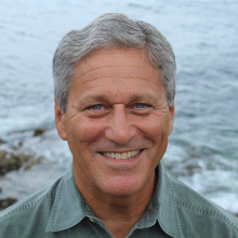 Male presenting person with short gray hair standing in front of the ocean.