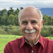Male presenting person in red flannel shirt standing in front of trees and mountains.