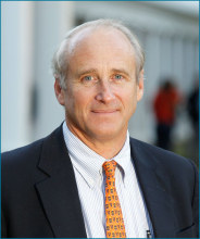 Male presenting person with short gray hair wearing a blue suit and orange tie.