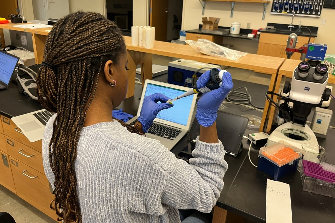 A student uses a pipette on a lab bench infront of a computer and microscope