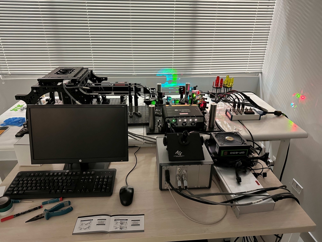 A computer monitor, keyboard, and mouse on a table along with microscopy equipment. An adjacent table positioned behind the first also has microscopy equipment on it.